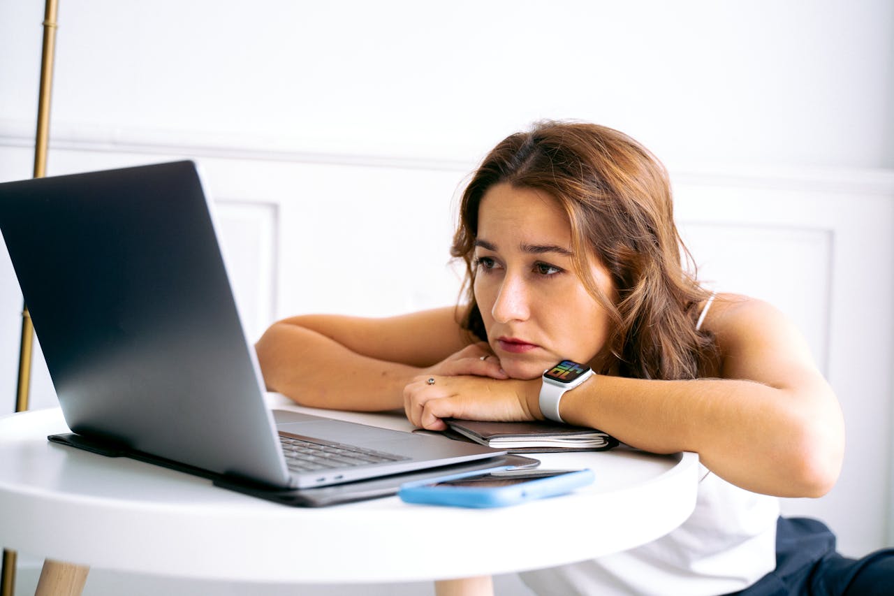 Woman appears tired while looking at a laptop, leaning on a table, in a minimalist indoor setting.
